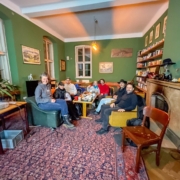 Medienstammtisch im Coconat – Dezember 2024 Gruppenfoto des Medienstammtischs im Coconat: mehrere Teilnehmer sitzen gemütlich im Salon zusammen, mit Beamer, Snacks und Bücherecke im Hintergrund.